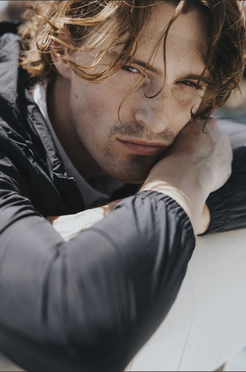 Close-up portrait of a male model resting his head on his arms, wearing a dark jacket outdoors.