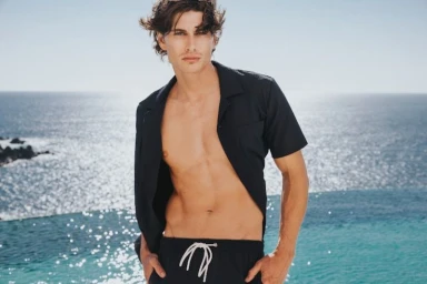Male model wearing an unbuttoned black short-sleeve shirt and black swim trunks outdoors against an ocean view