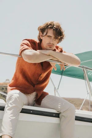 Male model in an orange shirt and white trousers leaning on a boat railing against a clear sky background