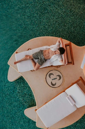 Aerial view of a male model relaxing on a wooden lounger by a turquoise swimming pool for the Scappino SS 2026 campaign.
