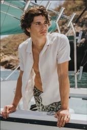 A young male model with dark curly hair wearing an unbuttoned white shirt and patterned swim shorts aboard a boat.