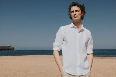 Male model wearing a white linen shirt posing on a sandy beach under a clear blue sky