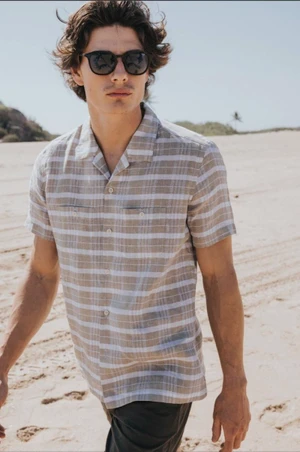 Male model wearing sunglasses and a striped short-sleeve shirt walking on a sandy beach.
