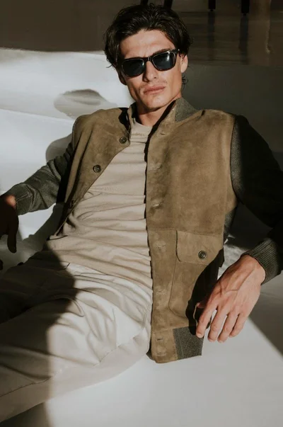 Male model wearing dark sunglasses, an olive suede jacket, beige t-shirt, and light trousers reclining on a white set.