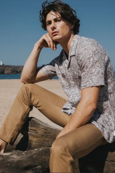 Male model wearing a patterned short-sleeve shirt and khaki trousers seated outdoors near the ocean