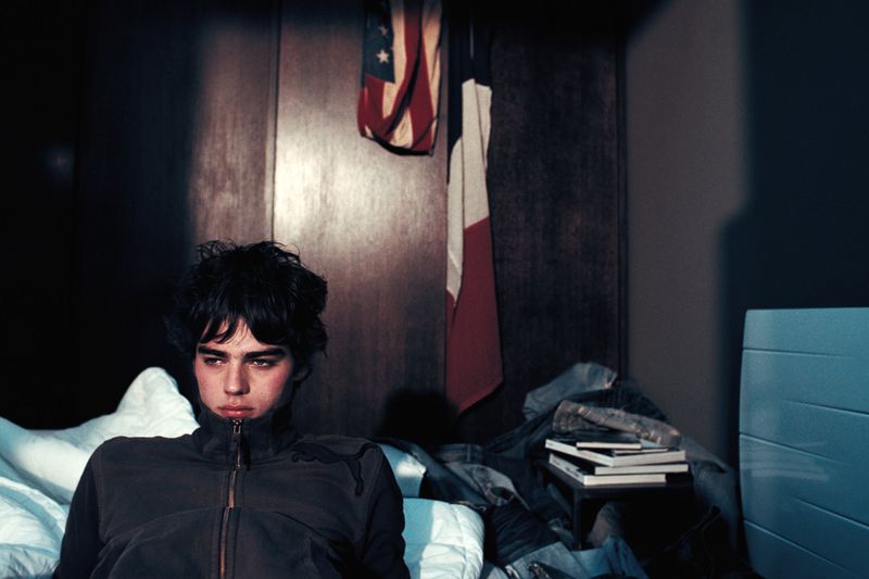 Young man in a dark zipped jacket reclining on a white bed with a hanging flag behind him