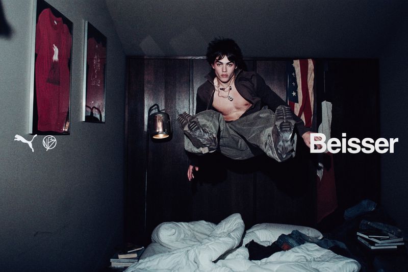 A young adult male mid-air wearing streetwear in a dark bedroom setting with an unmade bed, framed shirts on the wall, and warm lighting.