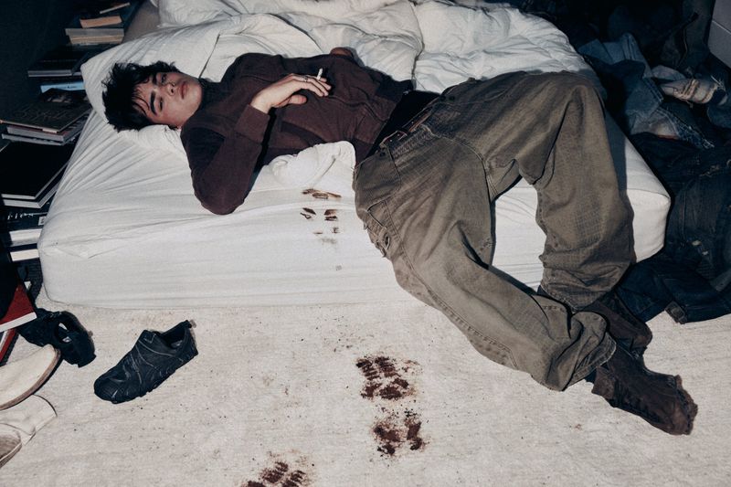 Young man lying on a white unmade bed wearing a brown top and loose grey trousers, with muddy shoe prints on the floor.