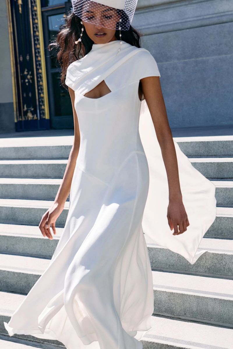 Model wearing a white bridal gown and netted pillbox hat walking down stone steps outdoors.