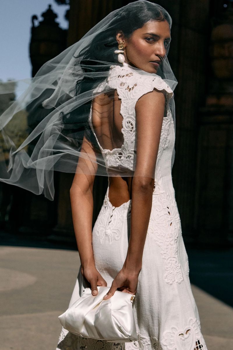 Model in a white lace bridal dress wearing a sheer veil and holding a small white pouch in an outdoor setting