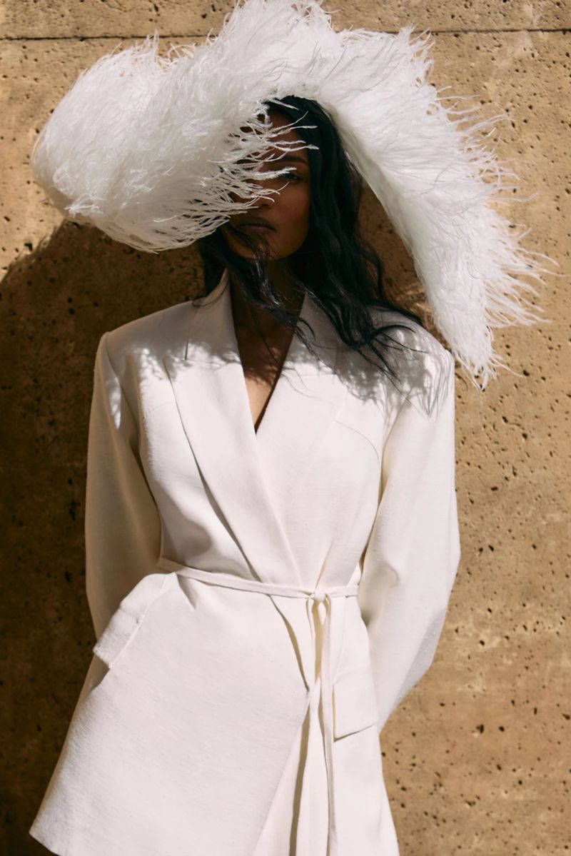 Model wearing a white blazer and large feathered hat against a textured stone wall in bright sunlight