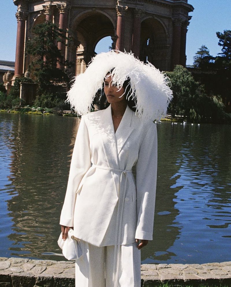 Model wearing a white suit and large feathery headpiece standing outdoors near a scenic lake with classical architecture