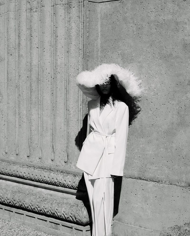 A woman stands against a textured gray wall wearing a white suit with a belted jacket and an oversized feathered hat in a monochrome image.