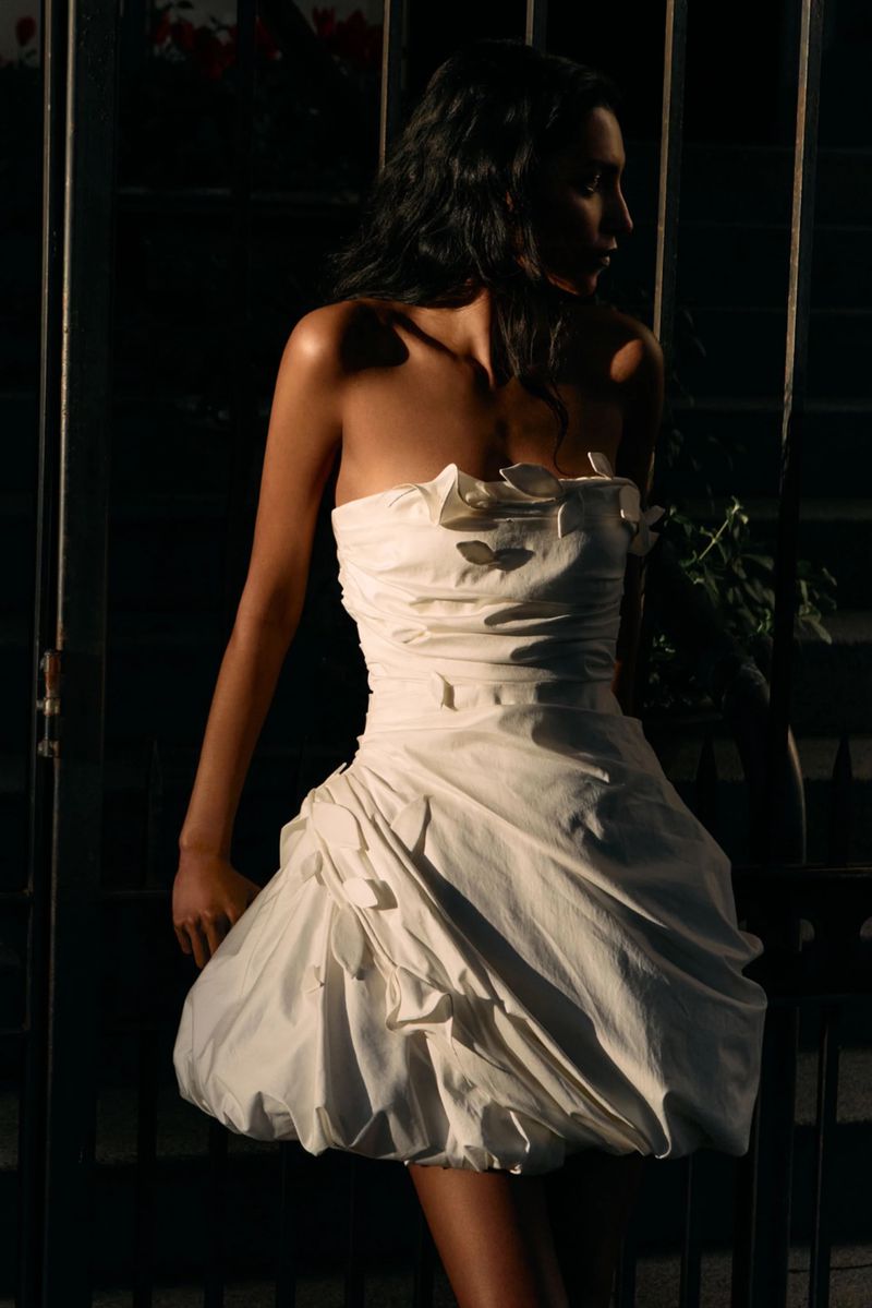 Model wearing a white strapless minidress standing against a dark metal gate in low-key dramatic lighting.
