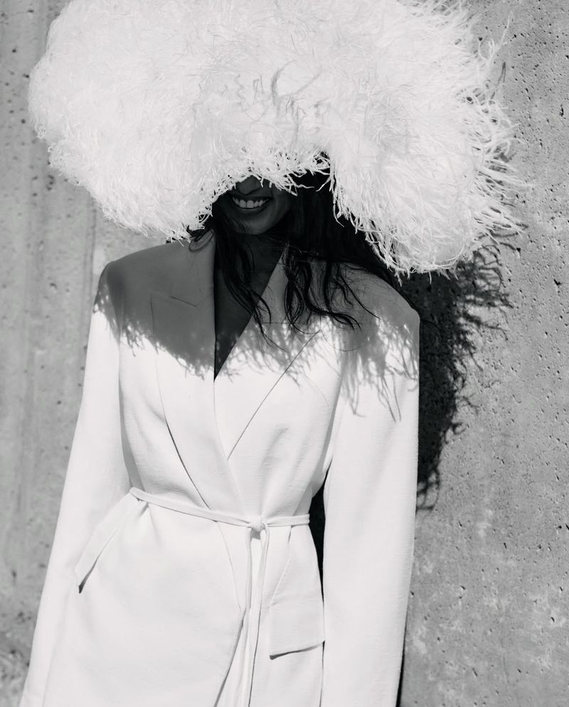 Model wearing a large white feather hat and belted jacket standing against a textured concrete wall.