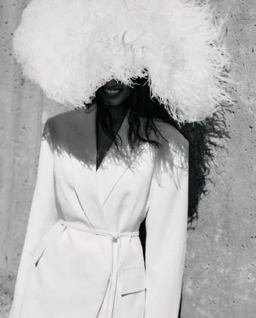 Model wearing a large white feather hat and belted jacket standing against a textured concrete wall.