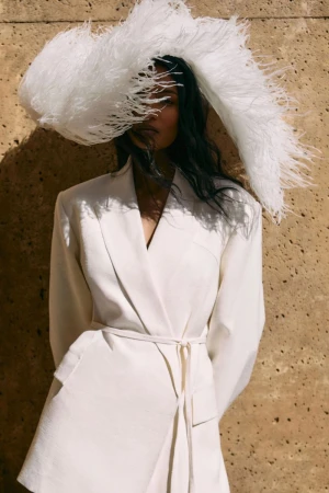 Model wearing a white blazer and large feathered hat against a textured stone wall in bright sunlight