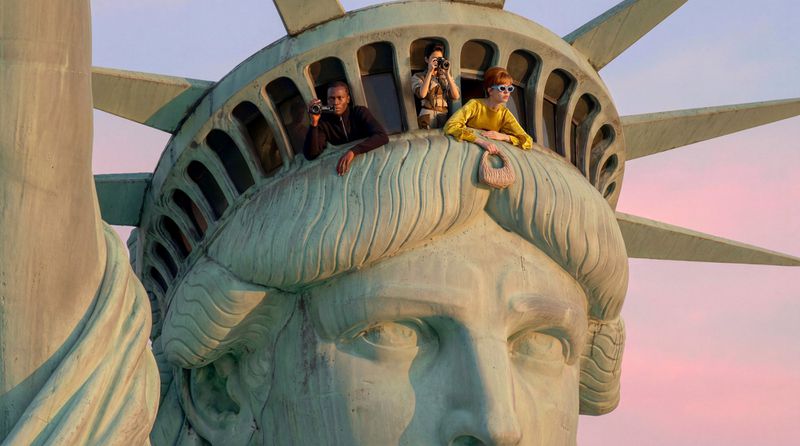 Three people leaning out from the crown windows of the Statue of Liberty during sunset.