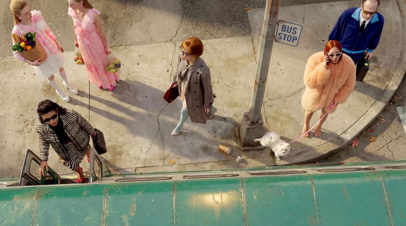 Eye-level, high-angle view showcases a diverse group of women and one man by a bus stop on an urban street with vintage flair.