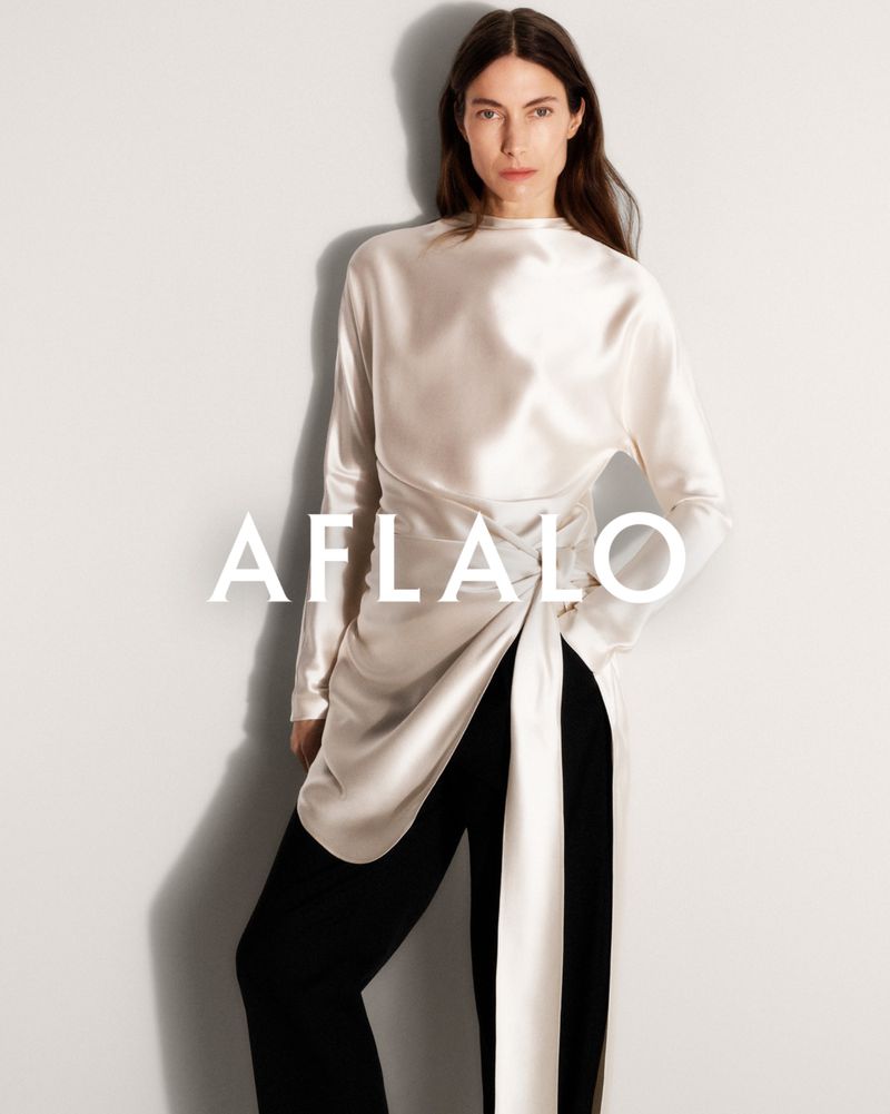Model Jessica Miller wearing a long white silk top and black trousers against a neutral white seamless studio background.