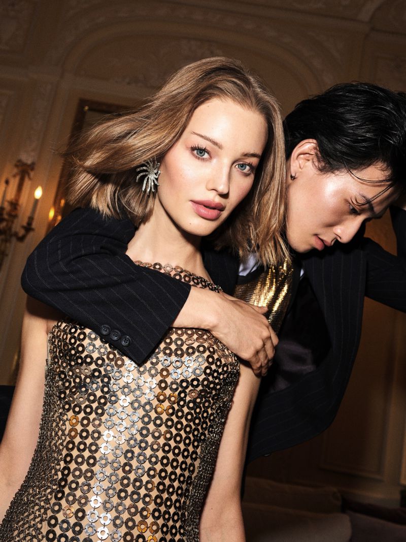 Two fashion models pose indoors: a fair-skinned woman in a metallic dress and a dark-haired man in a black suit with a gold shirt.