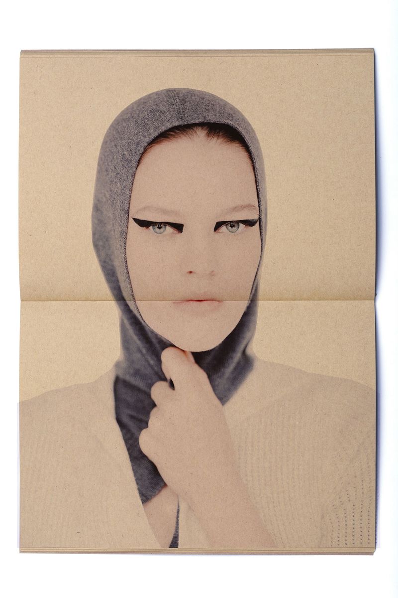 Close-up of a model wearing a grey hooded garment, heavy black eyeliner, and a cream-colored sweater, featured in a fashion editorial.