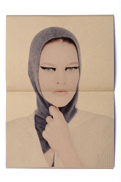 Close-up of a model wearing a grey hooded garment, heavy black eyeliner, and a cream-colored sweater, featured in a fashion editorial.