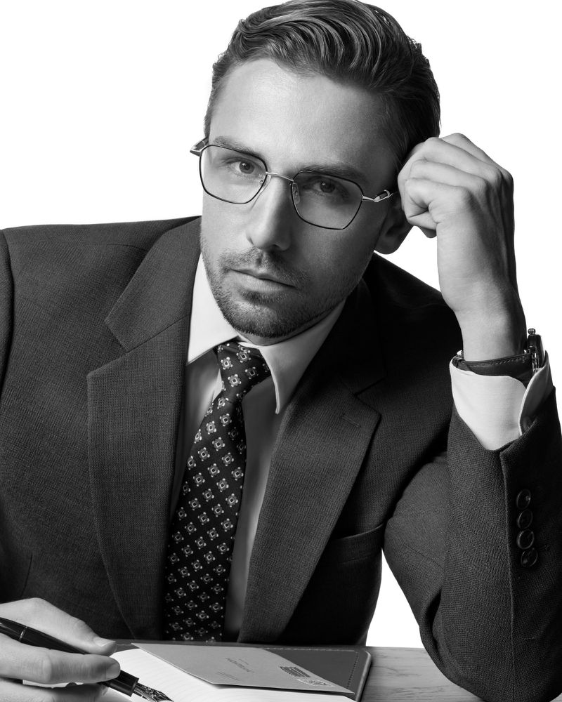 Black and white portrait of a pensive adult man in a suit and glasses, sitting at a desk with a pen and journal in a minimalist setting.