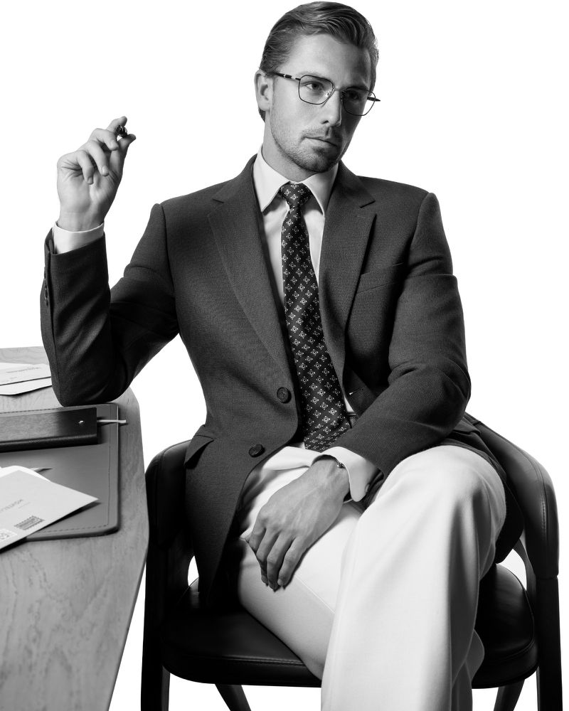 Man wearing eyeglasses, tailored suit, and tie seated at a desk against a white backdrop for the Montblanc Eyewear S/S 26 campaign.