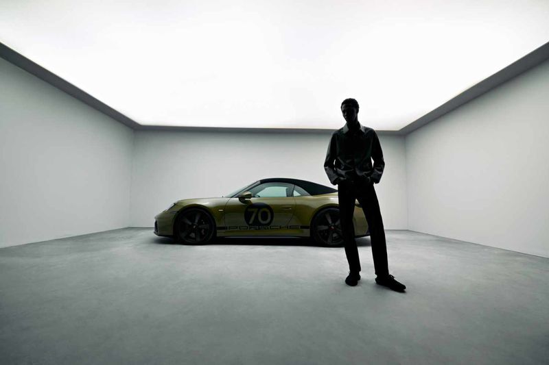 Model standing in silhouette next to a parked olive green sports car inside a white studio garage with a glowing overhead panel.