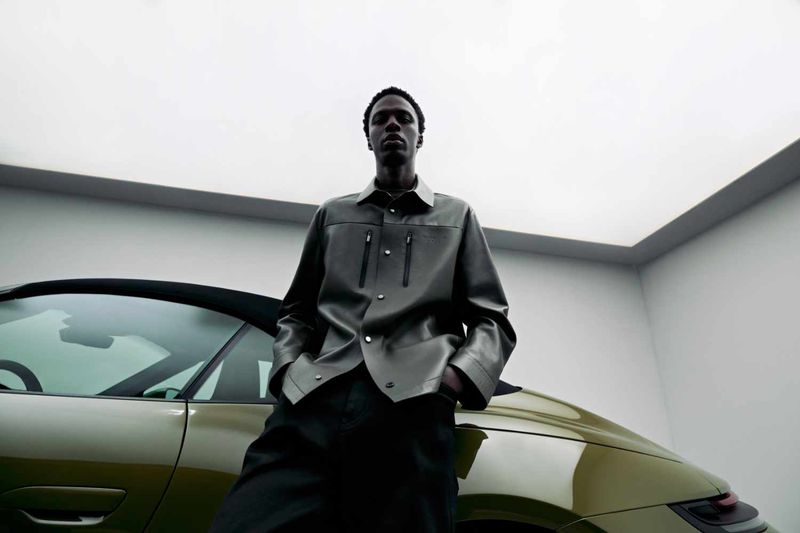 Model standing outdoors next to a luxury car under an overcast sky wearing a dark leather shirt and pants