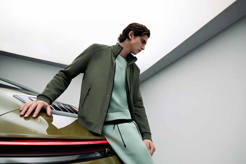 Model leaning against a green car wearing an olive jacket and light green sweatpants in a studio setting