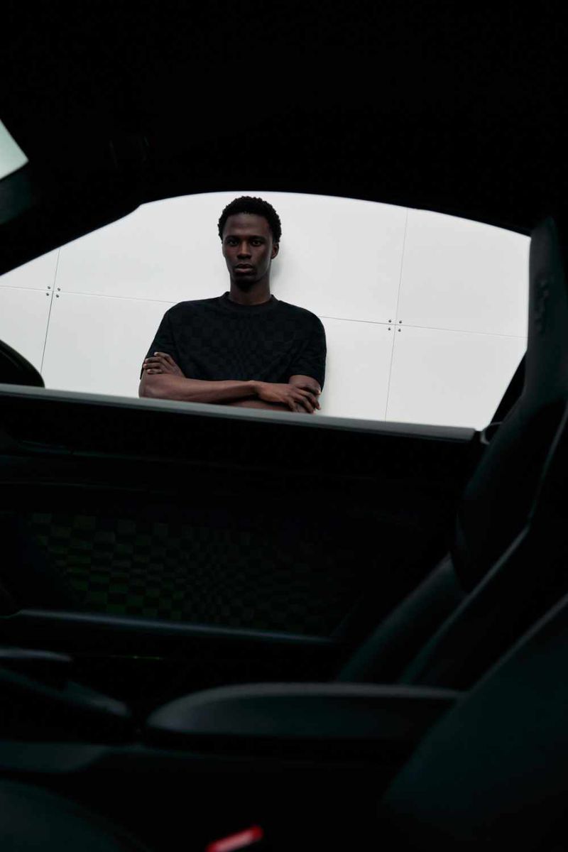 Indoor portrait of a dark-skinned adult male in a black T-shirt with arms crossed, inside an auto with visible black leather interior.