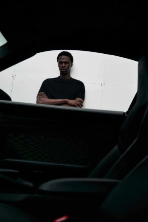Indoor portrait of a dark-skinned adult male in a black T-shirt with arms crossed, inside an auto with visible black leather interior.