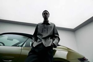 Model standing outdoors next to a luxury car under an overcast sky wearing a dark leather shirt and pants