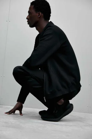 Black man crouching in profile wearing a black jacket and black trousers against a pale background