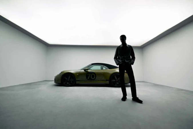 Model standing in silhouette next to a parked olive green sports car inside a white studio garage with a glowing overhead panel.