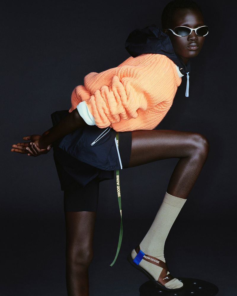 Fashion portrait of a Black woman wearing sunglasses, a salmon hoodie, and sandals, posing against a dark grey backdrop in a studio.