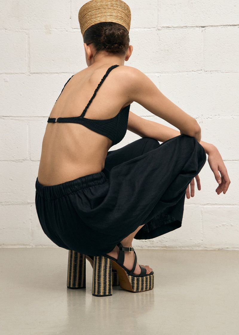 Model squatting in black crop top, dark trousers, and platform sandals against a white brick wall background