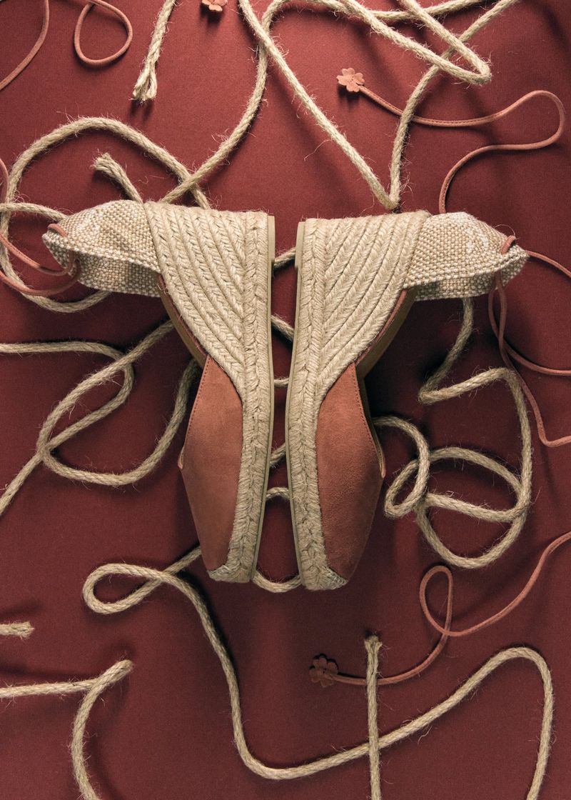 A pair of wedge espadrilles with woven jute heels and terracotta toes arranged symmetrically on a dark red surface with loose rope ties.