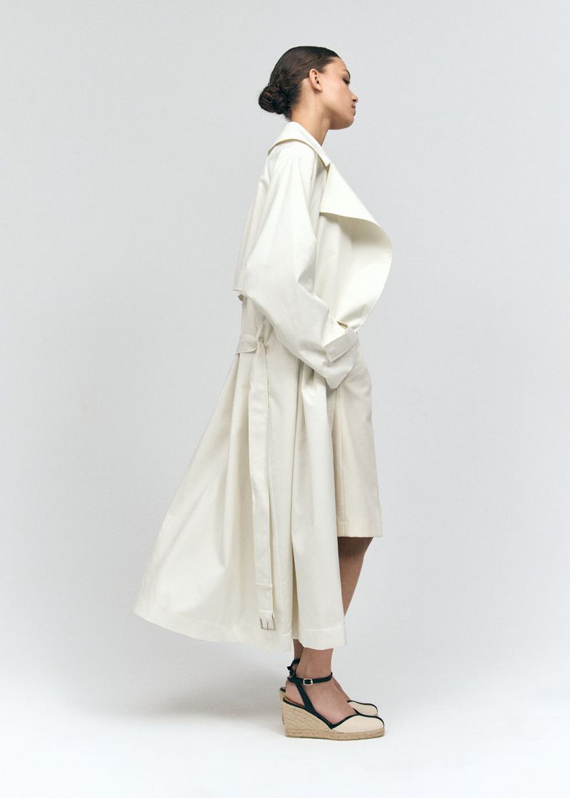 Model standing in profile wearing an ivory trench coat and espadrille wedge sandals against a white background