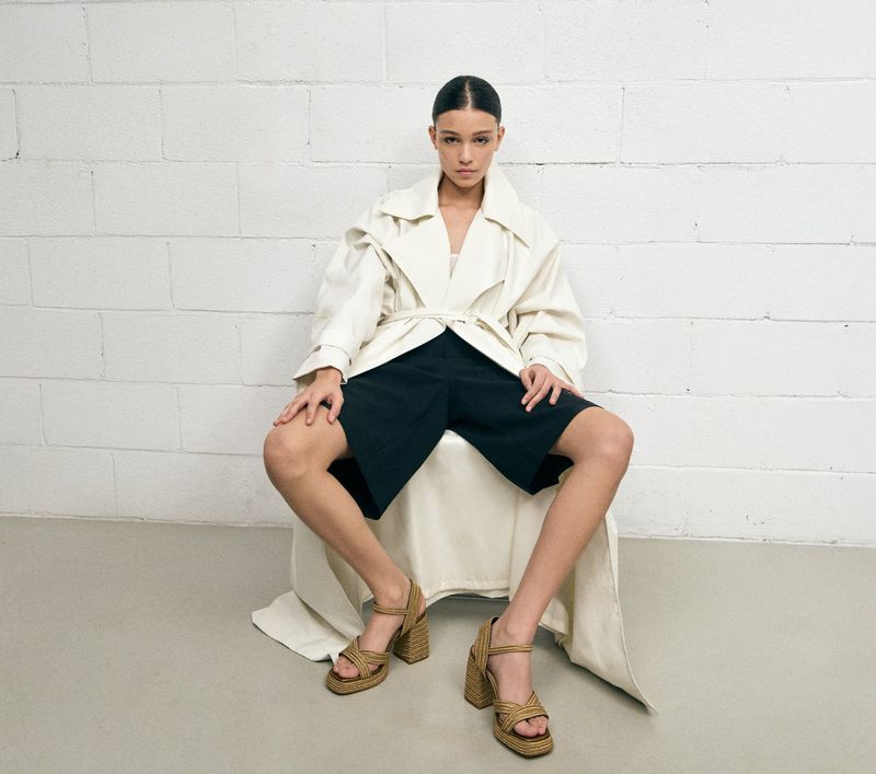 Model seated wearing an off-white trench coat, black shorts, and espadrille heels against a white brick wall