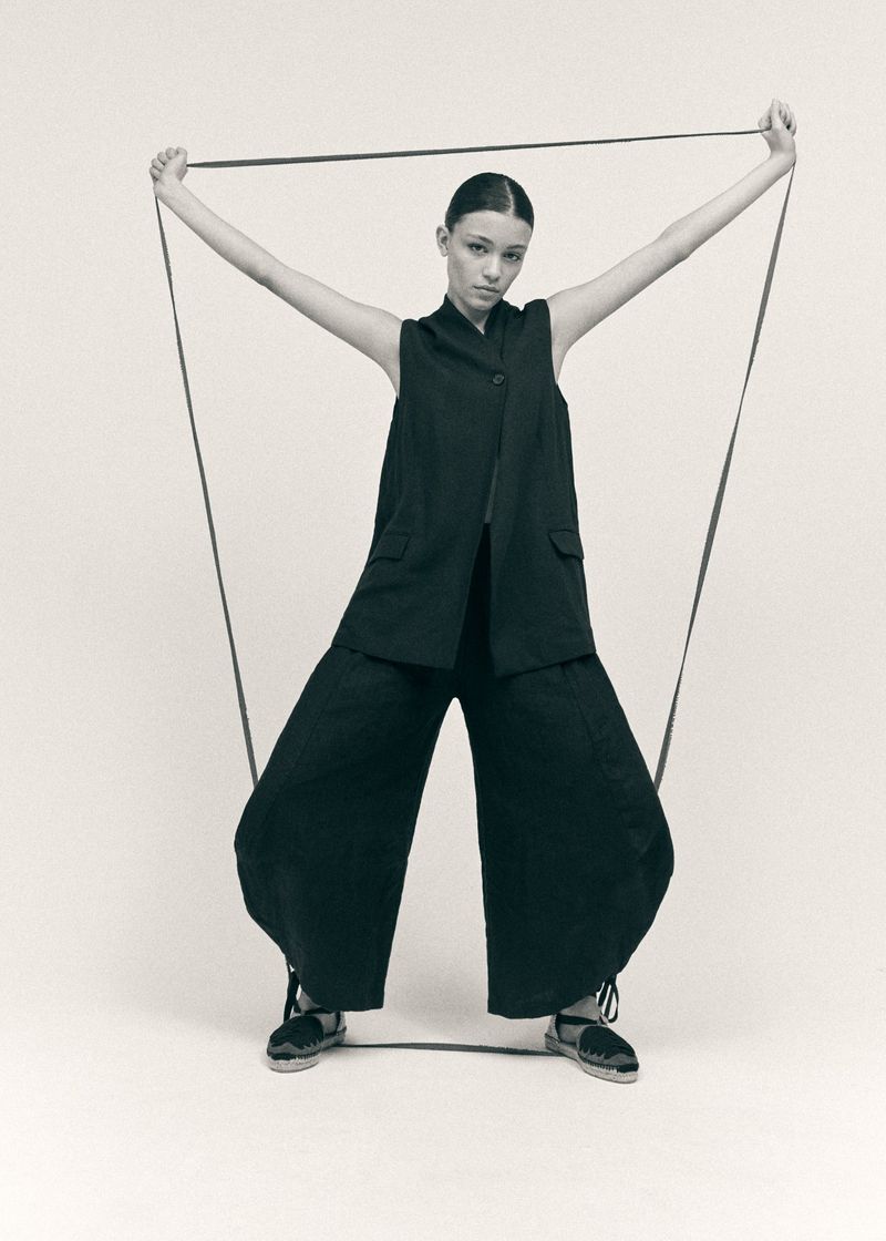 Model in black sleeveless vest and wide trousers holding straps against a plain backdrop for Castaner