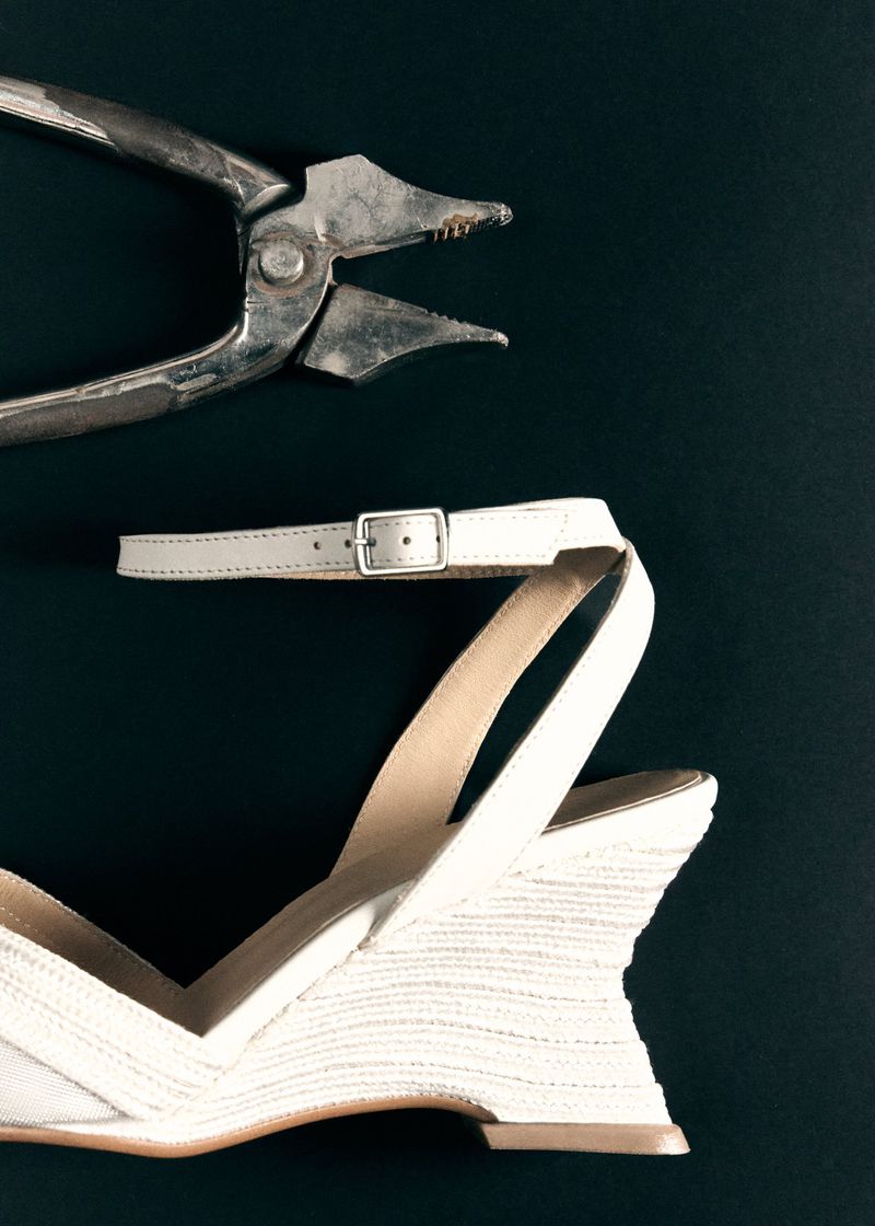 Close-up of a white wedge sandal with a buckled ankle strap and a pair of worn metal lasting pliers on a dark surface.