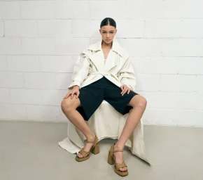 Model seated wearing an off-white trench coat, black shorts, and espadrille heels against a white brick wall