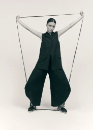 Model in black sleeveless vest and wide trousers holding straps against a plain backdrop for Castaner