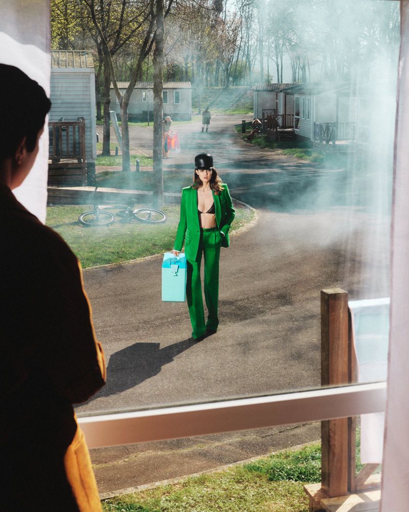 Lindsey Wixson in a green suit walking outdoors viewed through a window