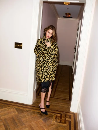 A woman in a patterned coat stands in a doorway of a hallway with a patterned wood floor and cream-colored walls in an indoor setting.