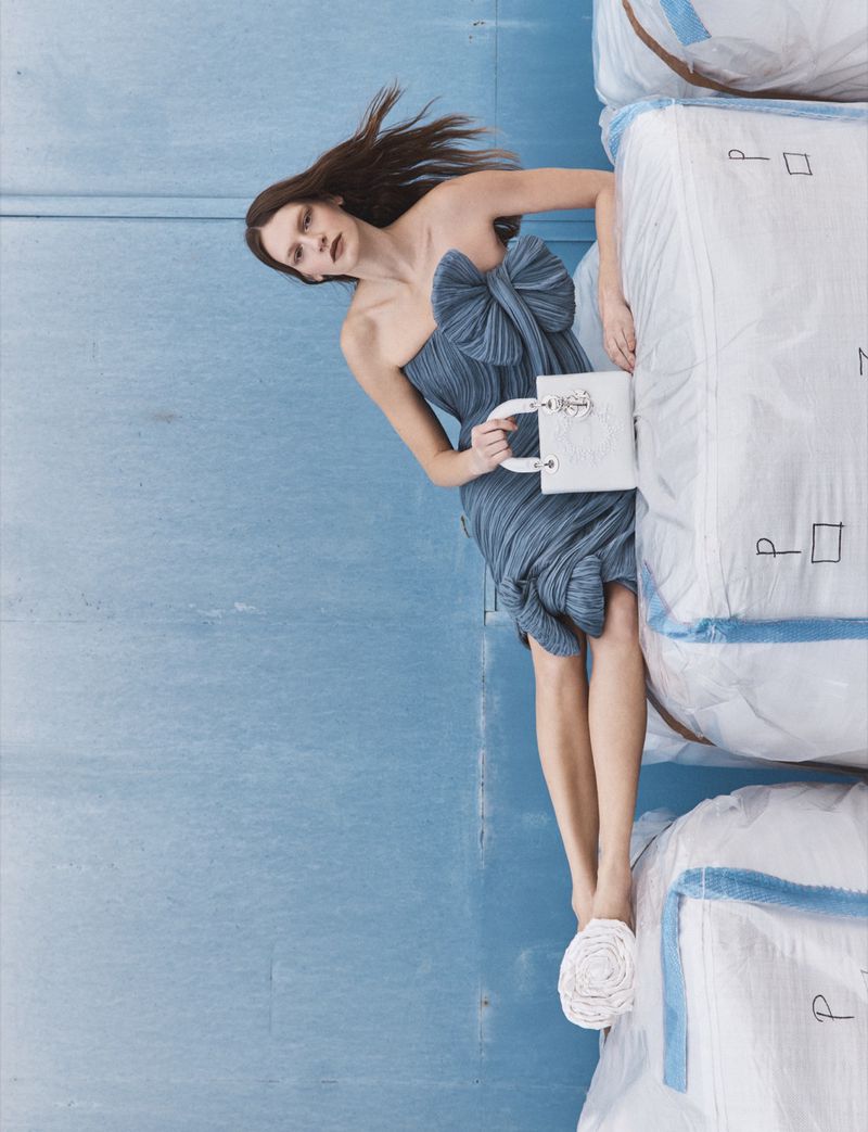 Model in strapless blue bow dress holding a white handbag against blue wall