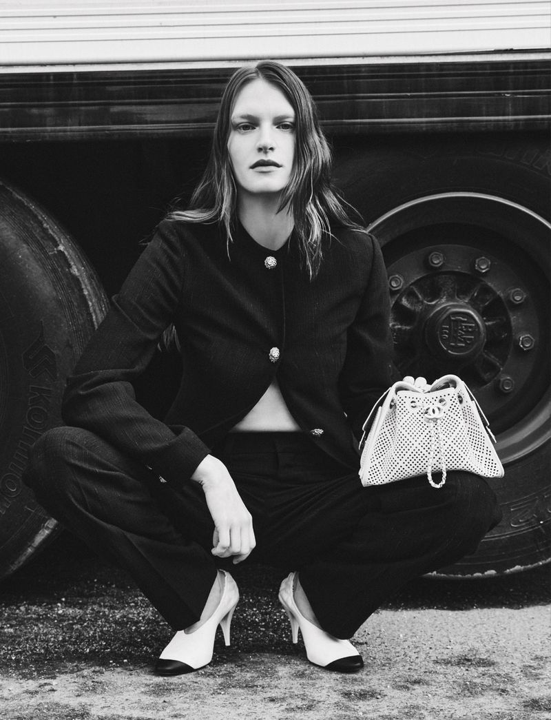 Model in dark jacket and trousers holding a patterned handbag while crouching in front of a truck tire